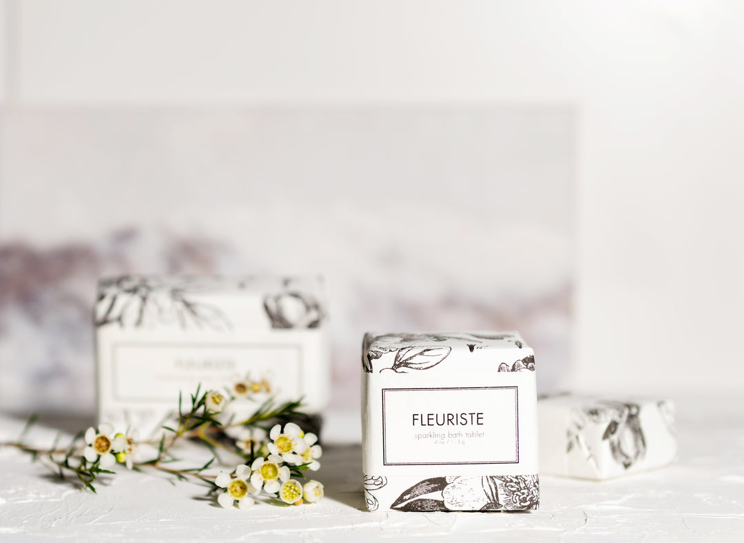 Two white soap bars labeled "Fleuriste" are on a white surface. The soap bars, decorated with floral illustrations, are accompanied by small white flowers. The background is blurred with soft, neutral tones, highlighting the products.