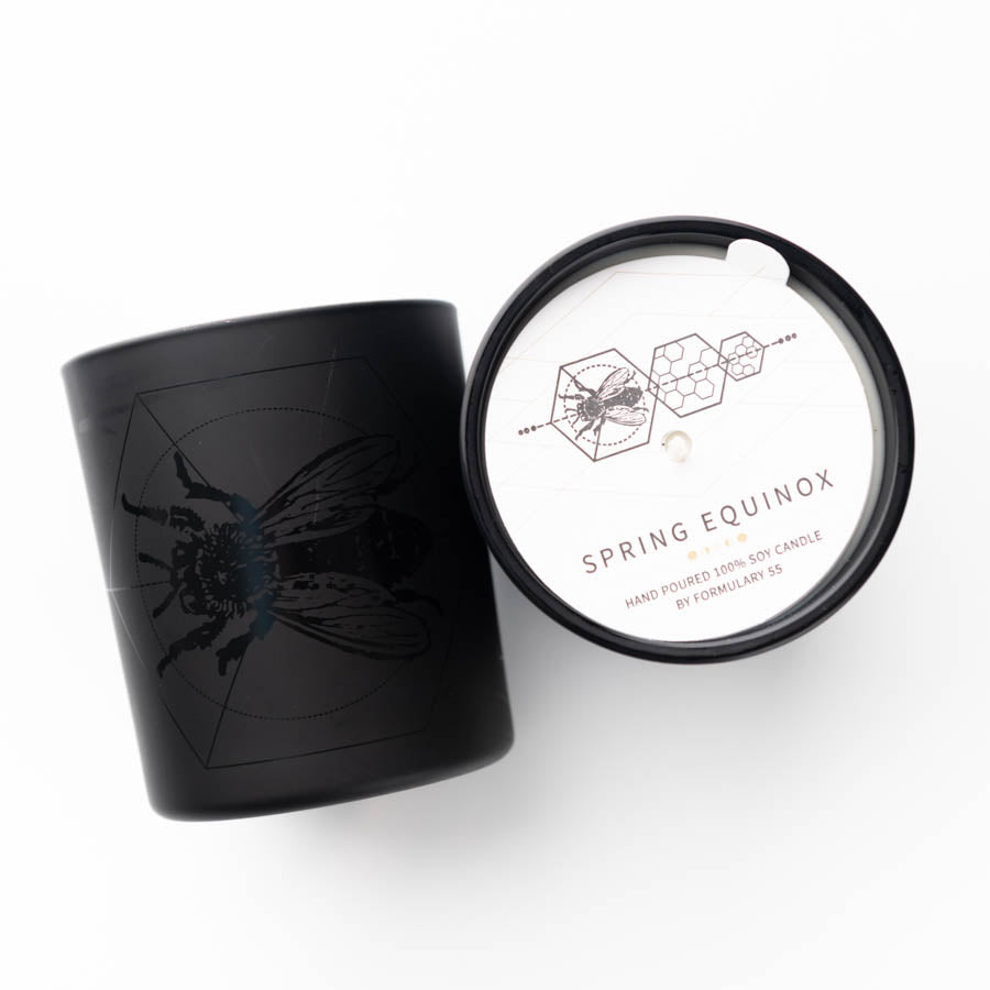 Black candle with an illustration of a bee on the side. The candle's lid is removed and lies next to it, showing a white label with geometric patterns, the bee illustration, and the words "Spring Equinox" and "Hand poured 100% soy candle by Formula J 59.