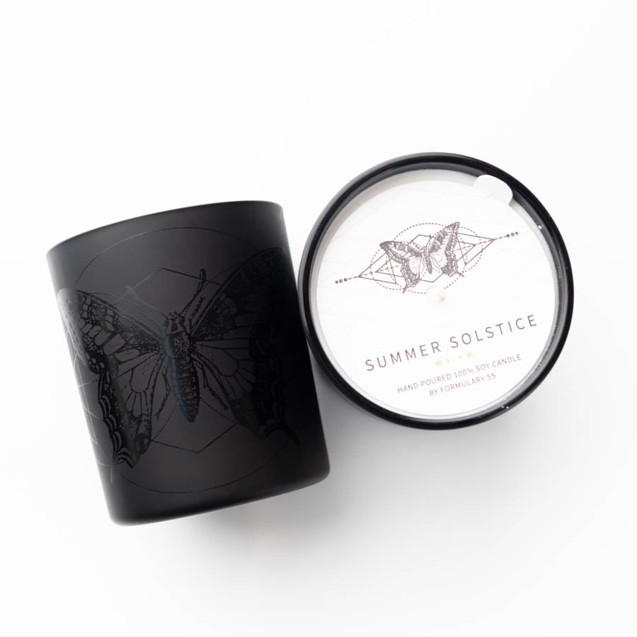 A black candle holder with a detailed moth design lies next to its lid, which is white and also features a moth illustration. The lid is labeled "SUMMER SOLSTICE" and notes that it's a hand-poured 100% soy candle by Formulary 55.