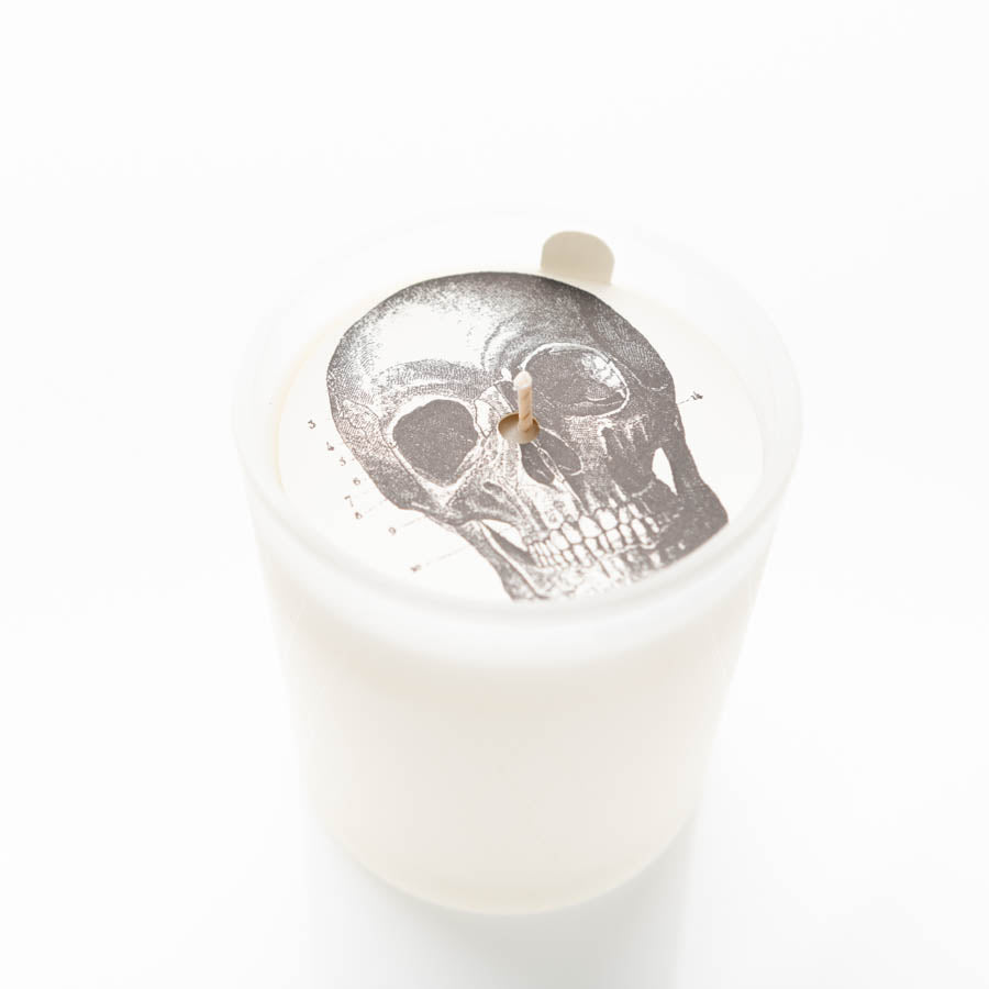 A white candle in a glass container. The candle has a printed image of a realistic human skull on the surface. The wick is unlit and centered in the middle of the skull image. The background is plain white.