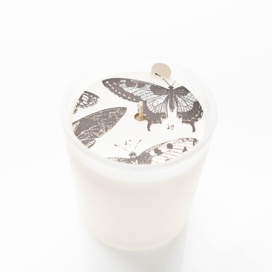 A white candle in a translucent white holder features a decorative cover with black-and-white drawings of butterflies. The wick is visible at the center of the candle, extending slightly from the cover.