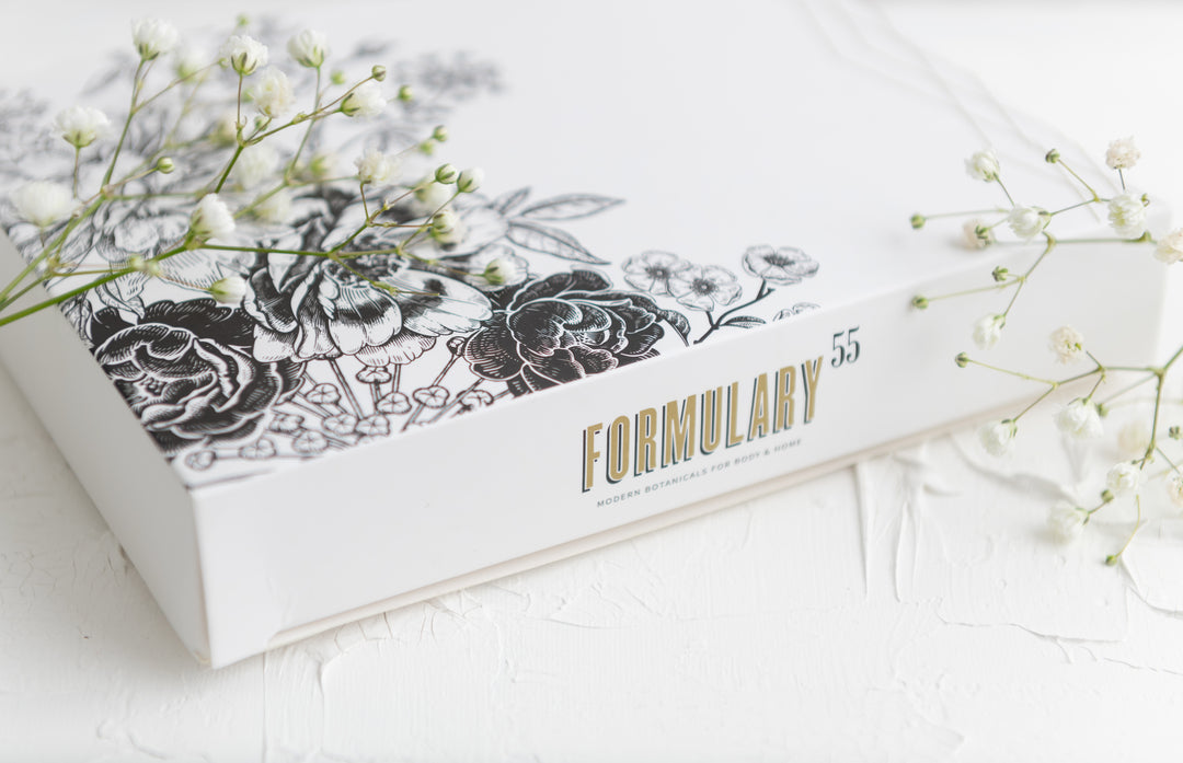 A white box labeled "Formulary 55" is adorned with intricate black floral illustrations. It is partially surrounded by delicate white baby's breath flowers, creating a soft, elegant atmosphere. The background features a textured white surface.