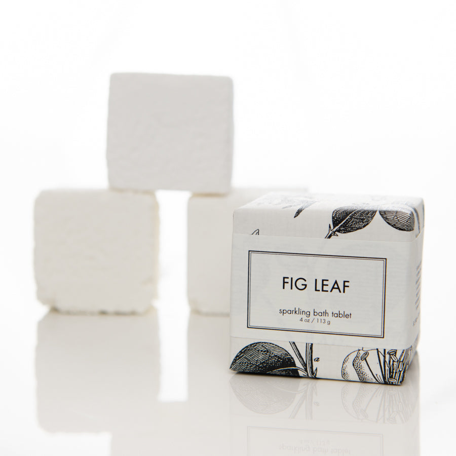 A close-up shot of a boxed product labeled "FIG LEAF Sparkling Bath Tablet" with a minimalist design featuring leaf illustrations. Three white bath tablets are stacked in the background on a clean, white surface.