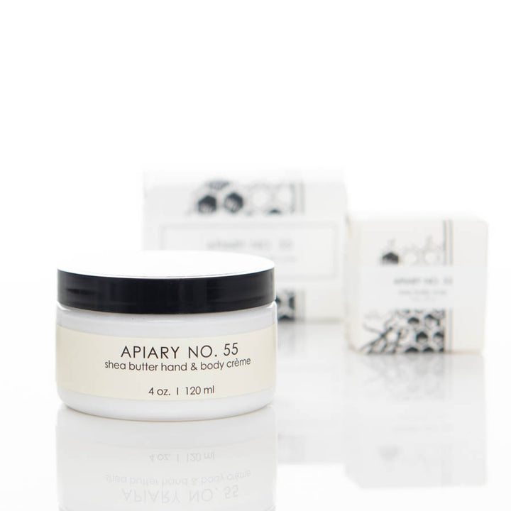 A 4-ounce container of APIARY NO. 55 shea butter hand & body crème is displayed in focus with a blurred background that includes the product's packaging. The container has a black lid and white label with black text.