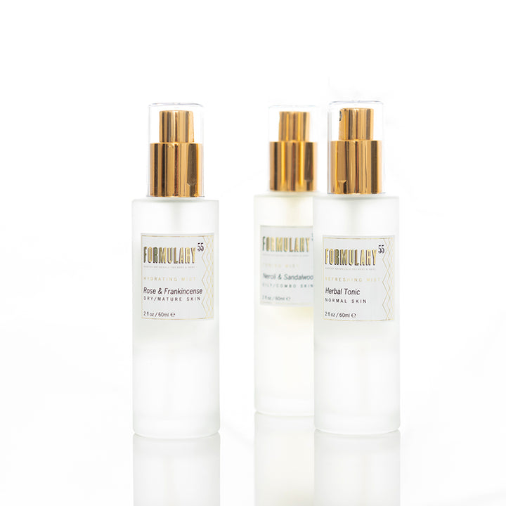 Three bottles of skincare products with gold-colored spray tops. The bottles are labeled "FORMULARY55" and include different formulations: "Rose & Frankincense," "Herbal & Sandalwood," and "Herbal Tonic." The bottles are arranged on a white surface.