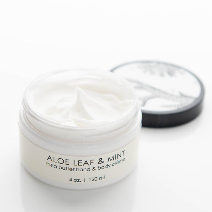 A white, open jar of Aloe Leaf & Mint shea butter hand and body crème, with a small white swirl of the cream visible. The black lid with a leaf design is placed in the background. The jar is labeled "4 oz. | 120 ml.