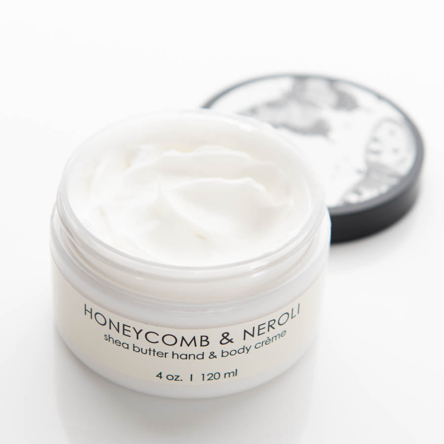 A white container with an opened lid showing a creamy white substance inside. The label reads "HONEYCOMB & NEROLI shea butter hand & body crème, 4 oz. | 120 ml." The lid, placed off to the side, has a black and white pattern.