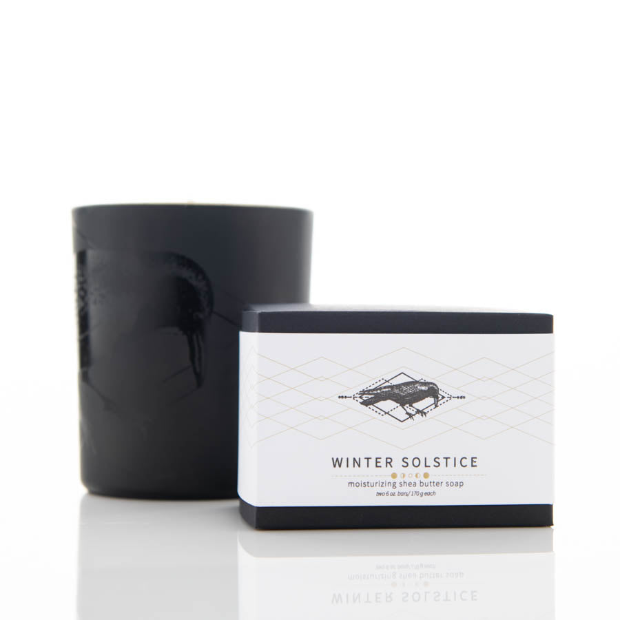 A black candle and a black and white box of "Winter Solstice" moisturizing shea butter soap are placed on a clean white surface. The candle has a subtle design, and the soap box features an elegant label with geometric patterns and text.