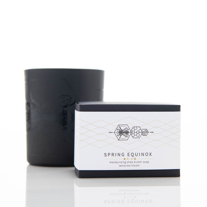 A bar soap packaged in a minimalist white and black box labeled "Spring Equinox" is placed beside a black candle holder. The soap box features geometric designs and the text indicates it is a moisturizing shea butter soap with beeswax and essential oils.