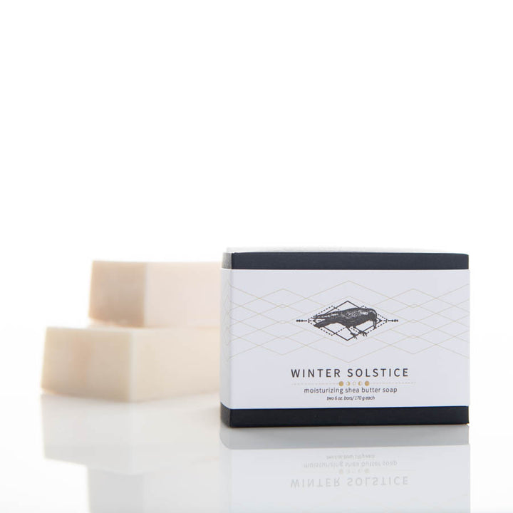 A box of Winter Solstice moisturizing shea butter soap is placed on a white surface with two unboxed bars of soap in the background. The packaging is minimalist, featuring a geometric pattern and text describing the product.