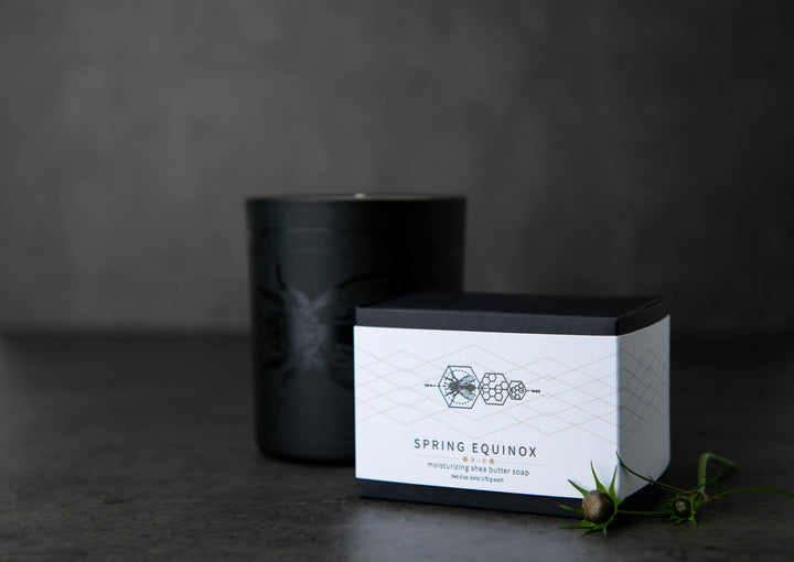 A black candle sits on a dark surface next to an elegant white box labeled "Spring Equinox," with a design featuring botanical illustrations. A sprig of green plant lies nearby, enhancing the minimalist and sophisticated presentation.