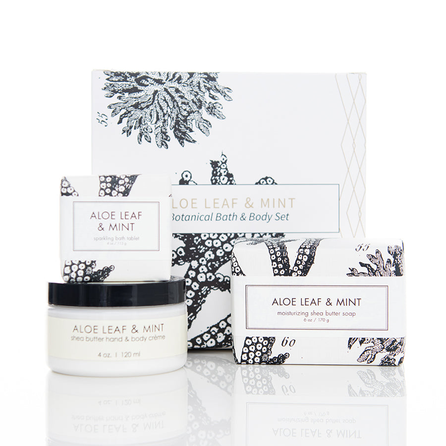 A botanical bath and body set with packaging labeled "Aloe Leaf & Mint." The set includes a shea butter hand and body crème jar, a shea butter soap bar, and a smaller shea butter soap bar. The packaging features black and white botanical illustrations.