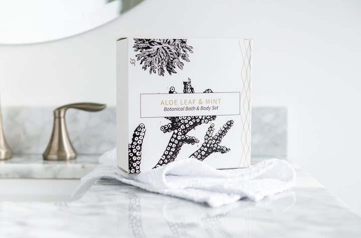 A decorative box labeled "Aloe Leaf & Mint Botanical Bath & Body Set" sits on a white marble bathroom countertop. The box features black and white botanical illustrations. A white towel and a silver faucet are also visible in the background.