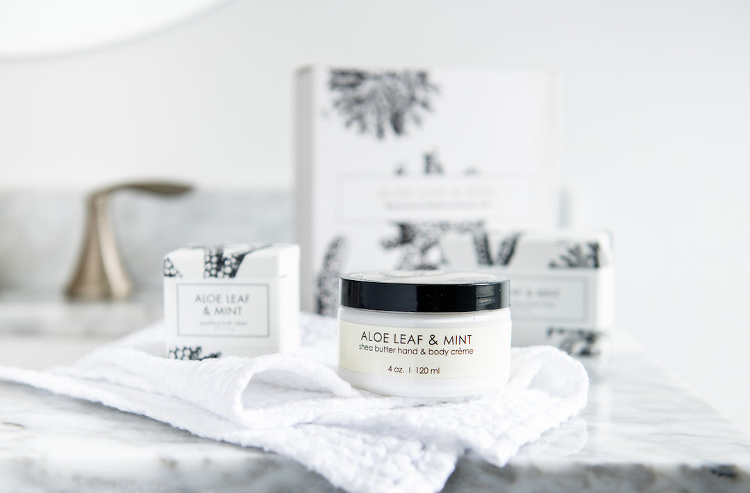 A jar labeled "Aloe Leaf & Mint Shea Butter Hand & Body Crème" sits on a white, textured towel on a marble surface. Background items include two product boxes with a similar design and a bathroom faucet in a blurred setting.