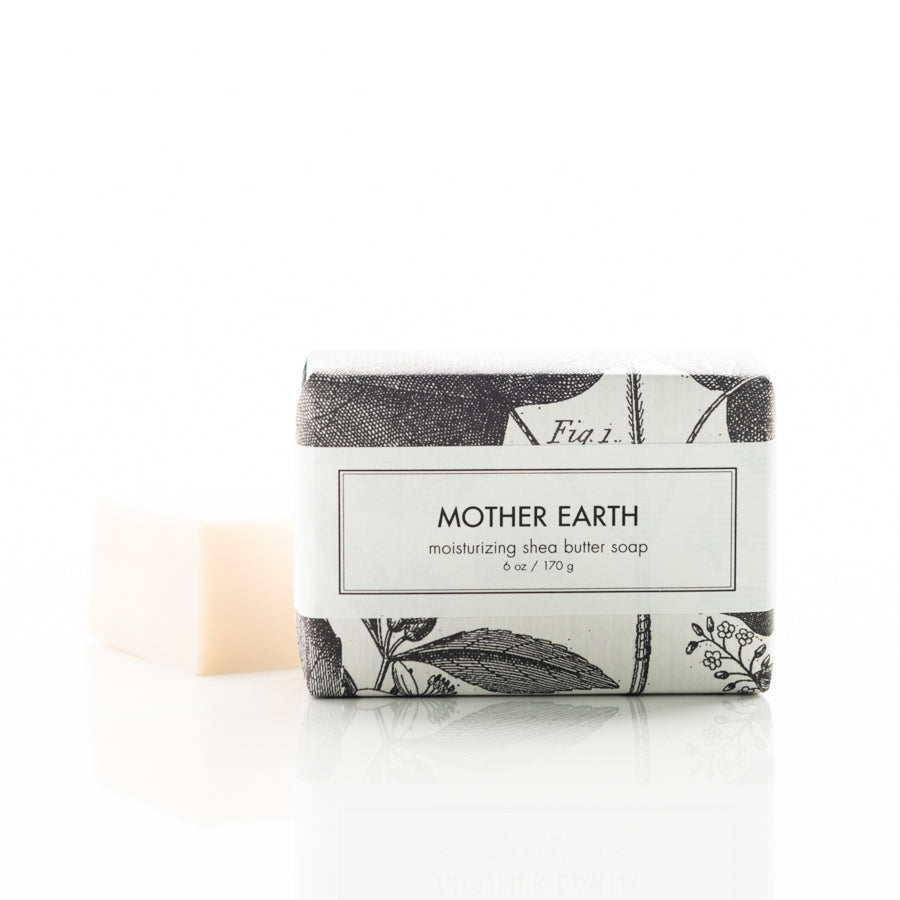 A wrapped bar of "Mother Earth" moisturizing shea butter soap is displayed with part of the unwrapped soap visible in the background. The packaging features elegant black botanical illustrations on a white background.