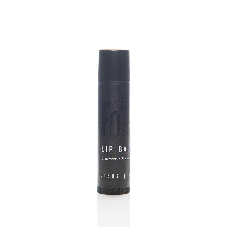 A black cylindrical tube of lip balm stands upright against a white background. The tube has minimalistic white text indicating it is a protective and moisturizing lip balm. The label also shows the weight, which is 0.15 oz.