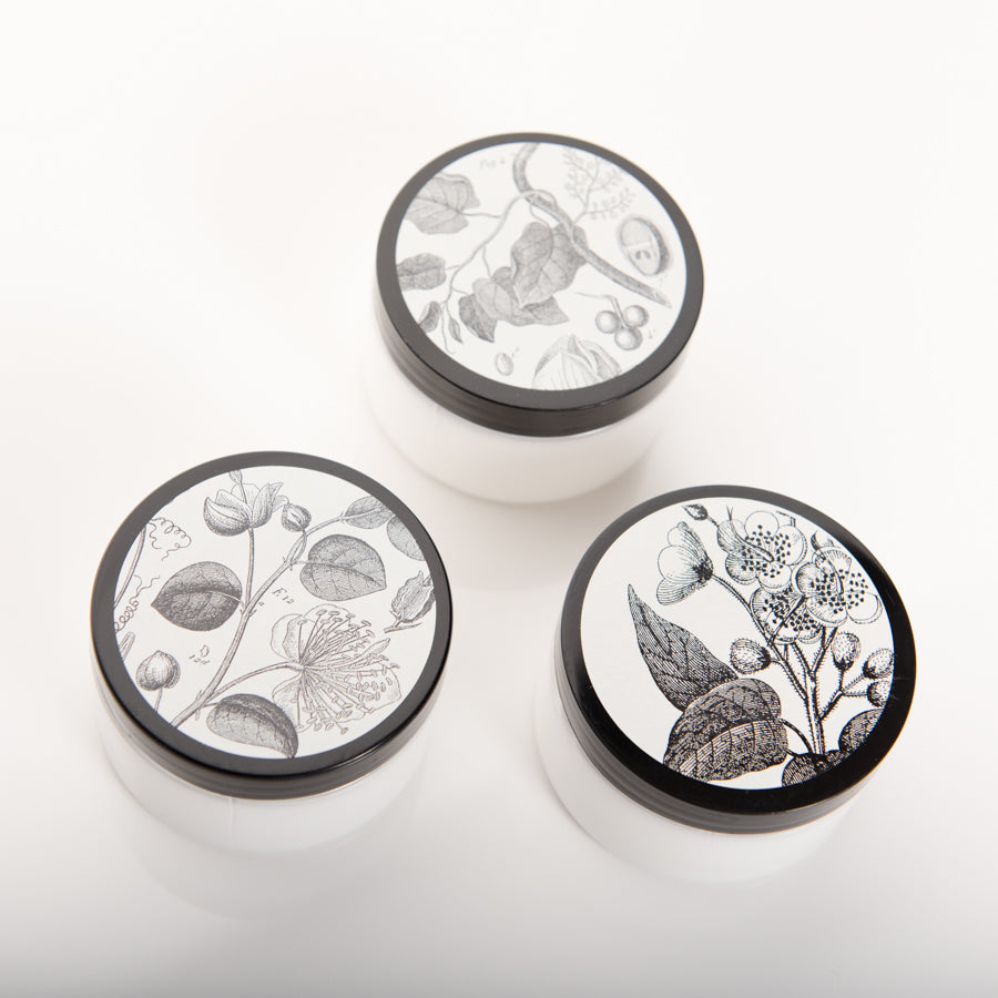 Three round white jars with black lids are arranged in a triangular formation. Each lid features a unique black and white botanical illustration, including various flowers and leaves, against a plain white background.