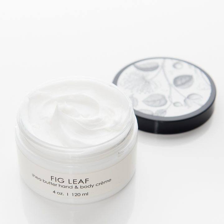 A white jar of Fig Leaf shea butter hand and body crème with a black and white illustrated lid displaying fig leaves and fruits. The jar is open, showing a smooth, creamy substance inside. The label indicates the size as 4 oz or 120 ml.