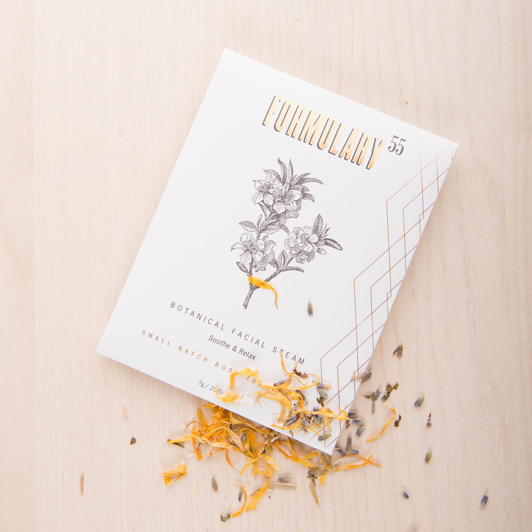 A package of Formulary 55 Botanical Facial Steam lies on a light wooden surface. The white packaging features golden text and a botanical illustration. Dried herbs and flower petals are scattered around and partially beneath the package.