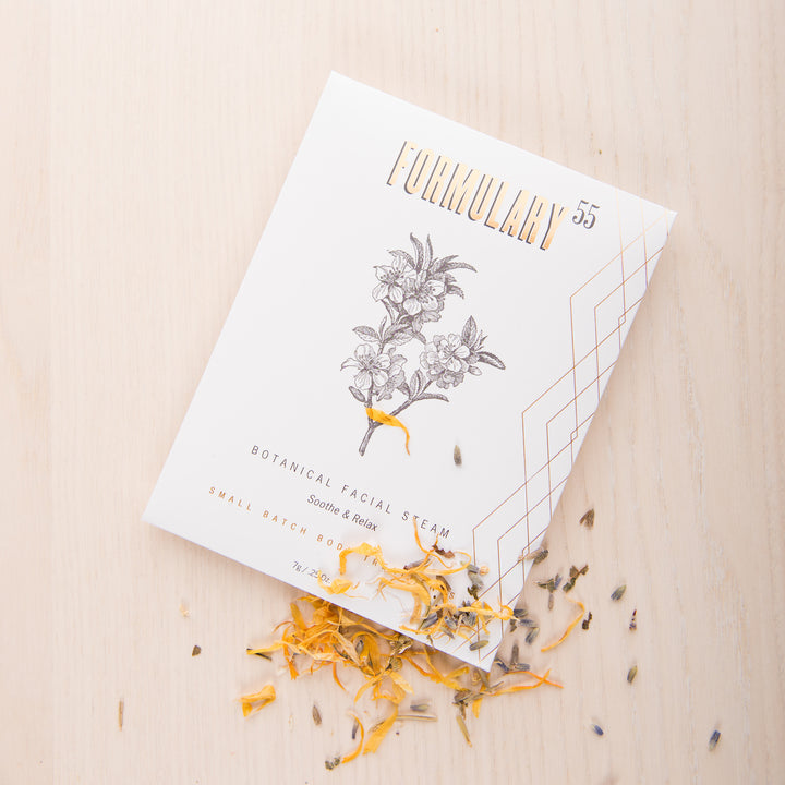 A package of Formulary 55 Botanical Facial Steam lies on a light wooden surface. The white packaging features golden text and a botanical illustration. Dried herbs and flower petals are scattered around and partially beneath the package.