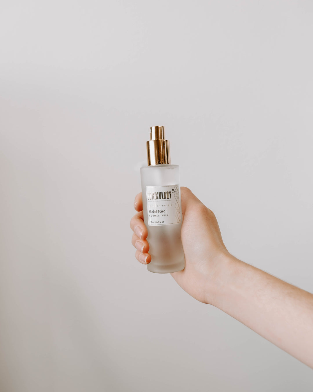 A hand holding a glass bottle of facial toner with a gold pump dispenser against a plain white background. The label on the bottle reads "The Ordinary." The hand is positioned such that the bottle is centered and clearly visible.