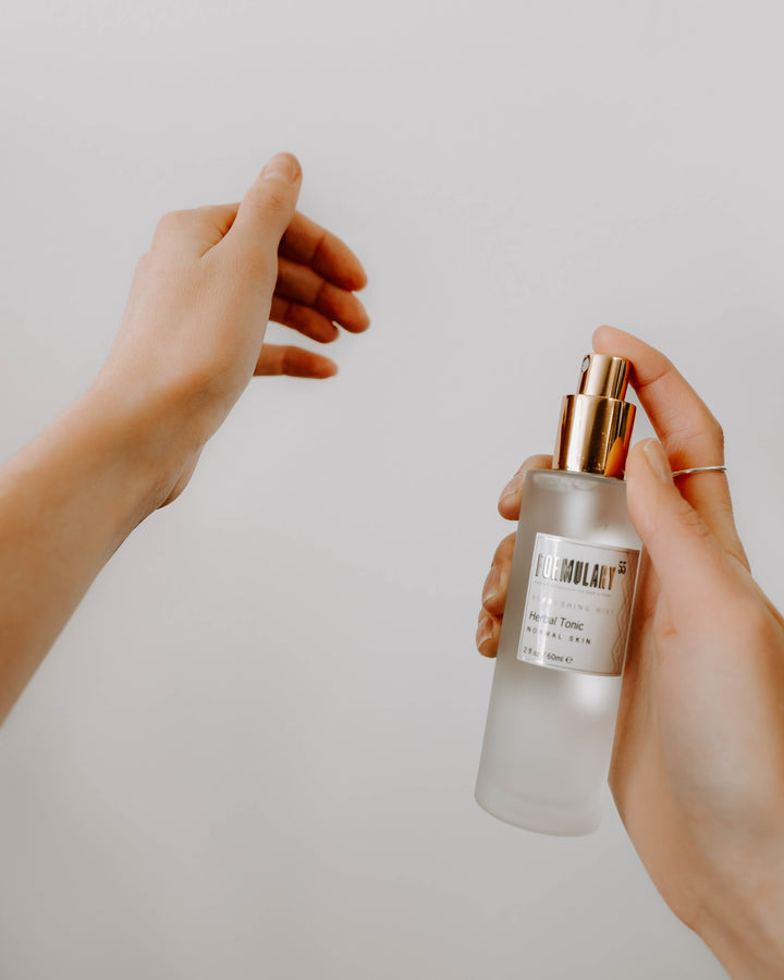 A person with light skin is holding a frosted glass bottle with a golden pump dispenser labeled "Formulati" in one hand while reaching out with their other hand. The background is plain and white.