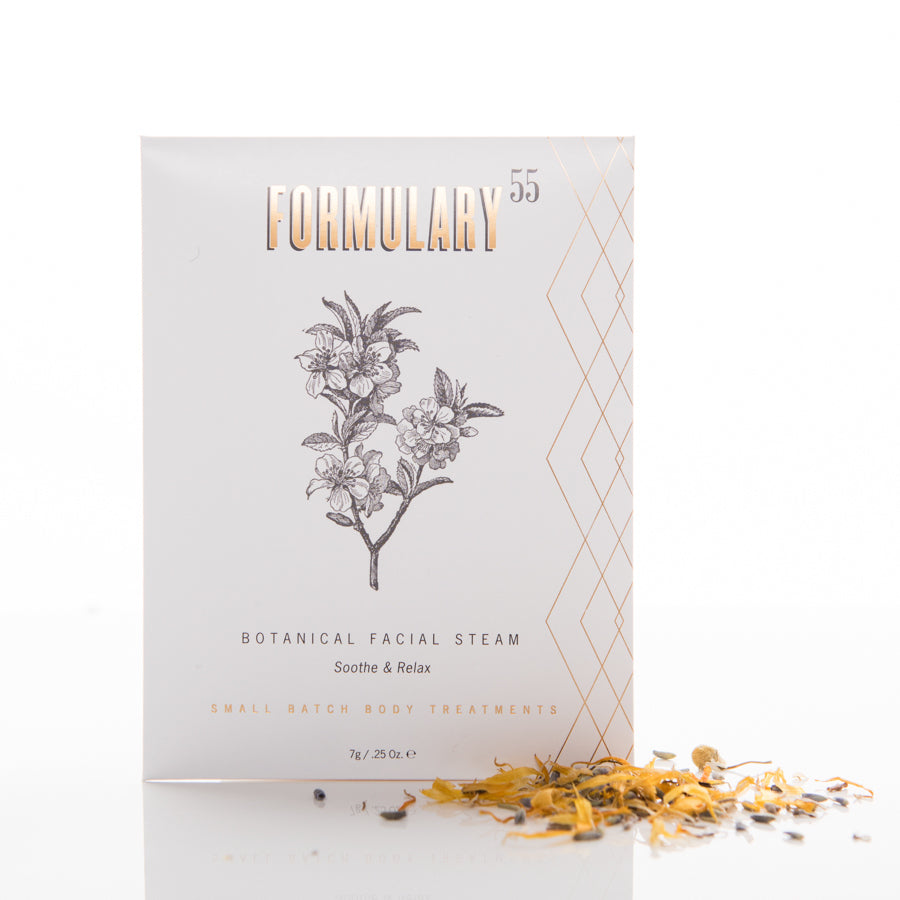 A white product box labeled "FORMULARY 55" with a botanical illustration on the front containing “Botanical Facial Steam - Soothe & Relax" and "Small Batch Body Treatments". Loose dried herbs are scattered near the box on a white surface.