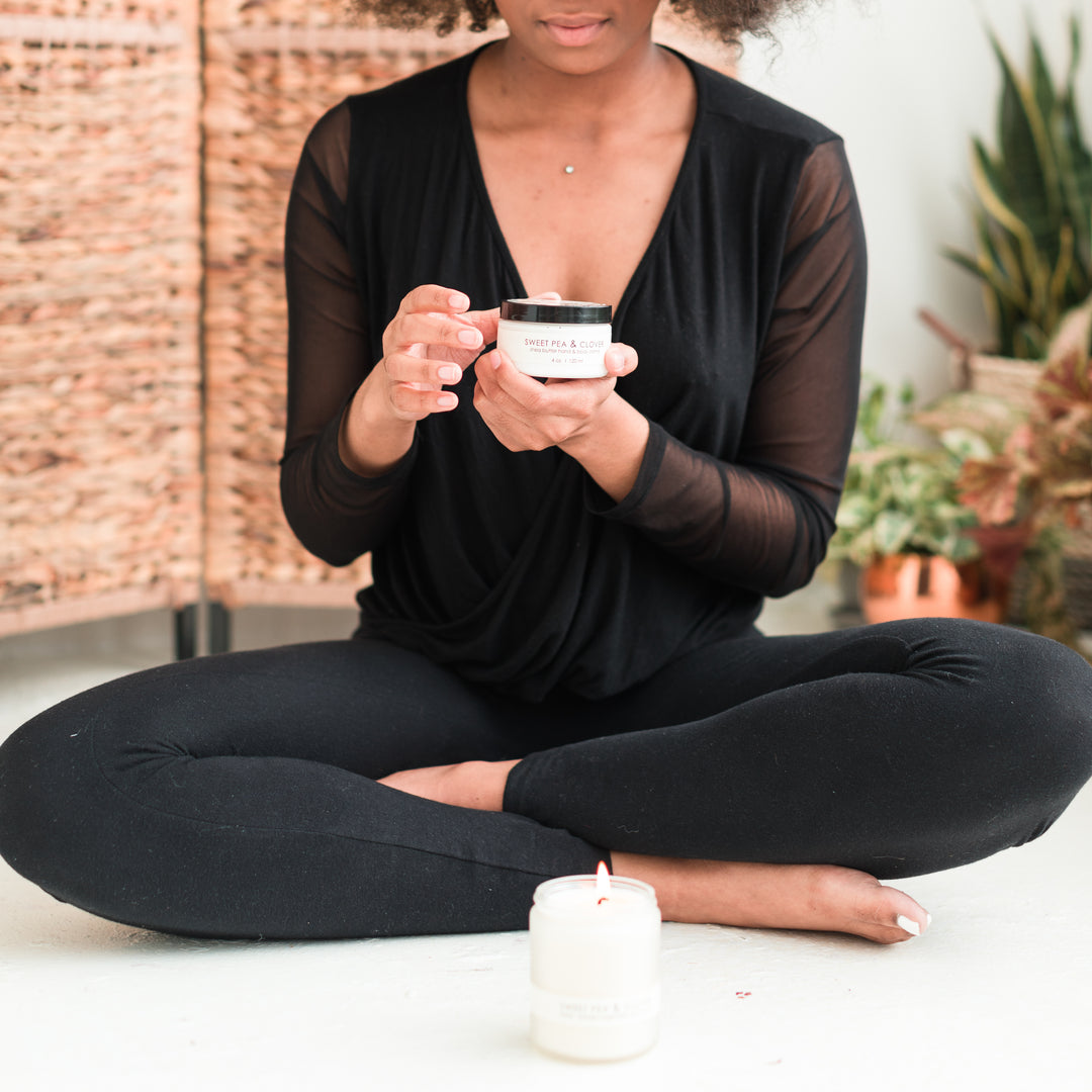 A person in black clothing sits cross-legged on the floor, holding a small white jar. A lit white candle is placed on the floor in front of them. Wicker furniture and potted plants are in the background.