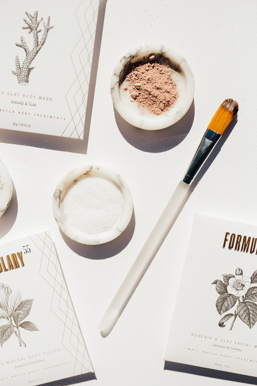 The image features skincare products on a white background: a round bowl with pink clay, a smaller bowl with white powder, a brush with a wooden handle, and packaging for a clay body mask and facial mask. The packaging is decorated with botanical illustrations.