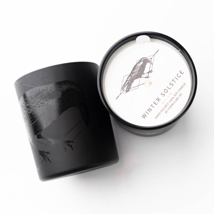 Black candle holder with a crow illustration. Lid removed, revealing a wick and label that reads "Winter Solstice" with further text "Raven Scented Soy Candle." The lid also features a similar crow illustration on a white background. Minimalist design.