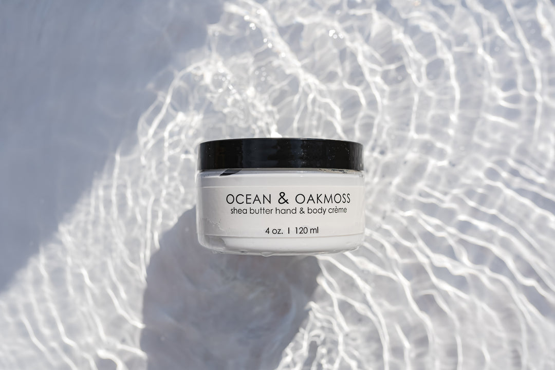 A jar of Ocean & Oakmoss shea butter hand and body crème, 4 oz. (120 ml), sits in shimmering, rippling water, creating a soothing and refreshing backdrop. The jar has a black lid and simple white label with black text.