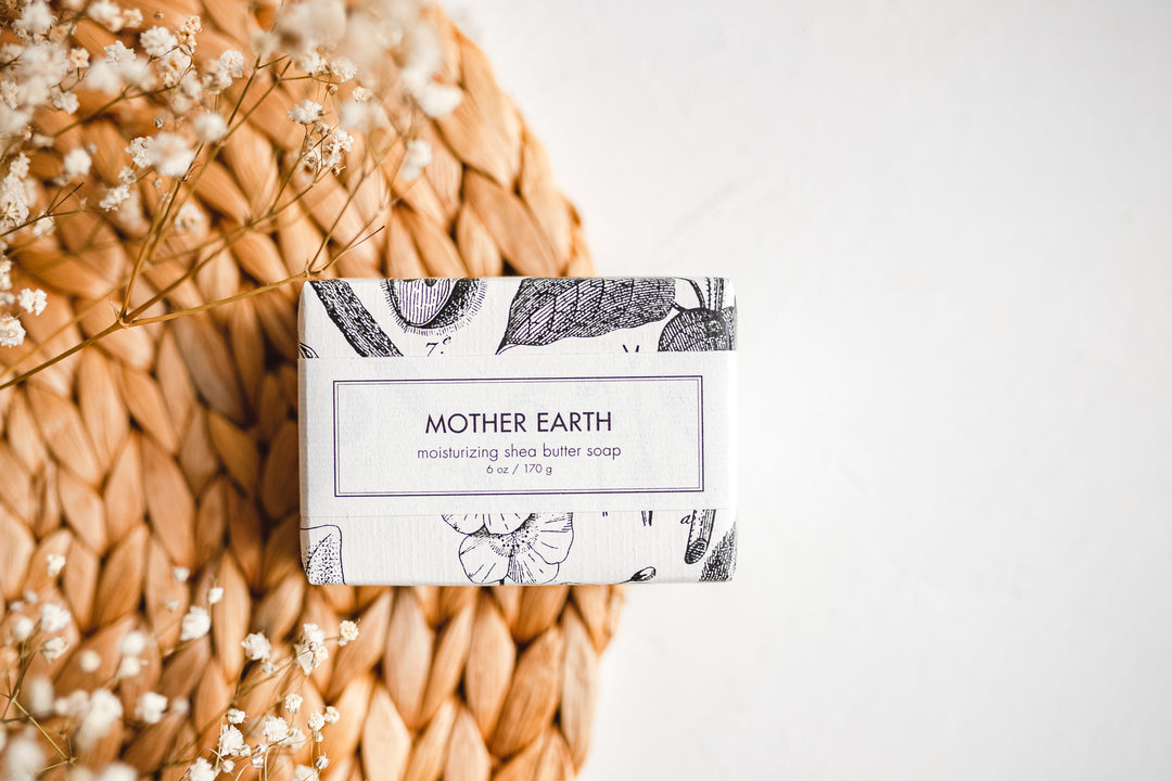 A rectangular bar of "Mother Earth" moisturizing shea butter soap is placed on a woven placemat with sprigs of white flowers next to it. The soap's packaging features botanical illustrations and text indicating it weighs 6 oz / 170g.