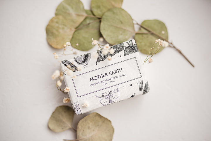 Bar of moisturizing shea butter soap labeled "Mother Earth," wrapped in floral paper with green and dried botanical leaves in the background.