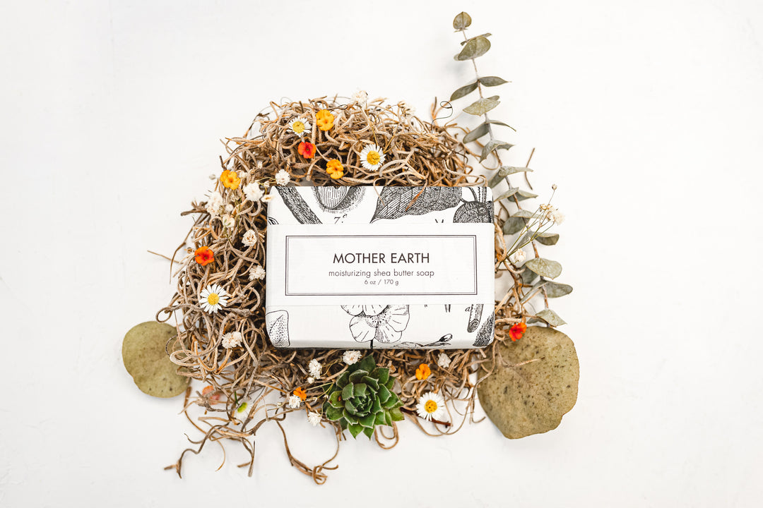 A bar of "Mother Earth" moisturizing shea butter soap is wrapped in white paper with black illustrations. It is surrounded by dried flowers, twigs, and leaves on a white background. Two stones and a succulent plant are also arranged around the soap.