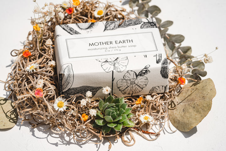 A box of "Mother Earth" moisturizing shea butter soap wrapped in illustrated paper with floral and leaf designs. The box is placed in the center of a decorative arrangement of dried flowers, leaves, and a succulent plant on a white background.