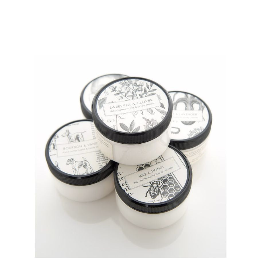 Five small jars of hand cream stacked together. Each jar has a different label design, including "Sweet Pea & Clover", "English Lavender", "Bourbon & Vanilla", and "Milk & Honey". The jars have white bases and black lids with illustrated labels.